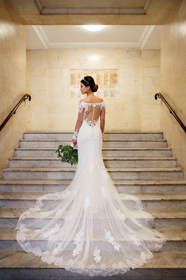 A bride in a stunningly beautiful full-length wedding dress with a lace train, poses on the marble steps of The Old Marylebone Town Hall.