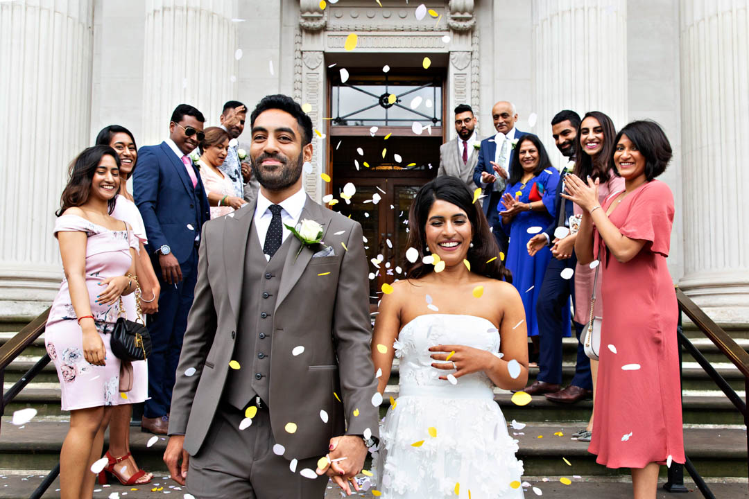 A bride and groom smile as they are covered in paper confetti by their families.