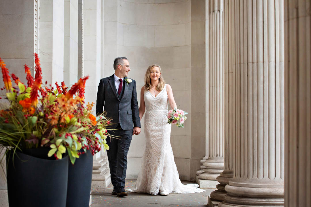 A bride and groom walk along the columns of Old Marylebone Town Hall, holding hands and smiling.