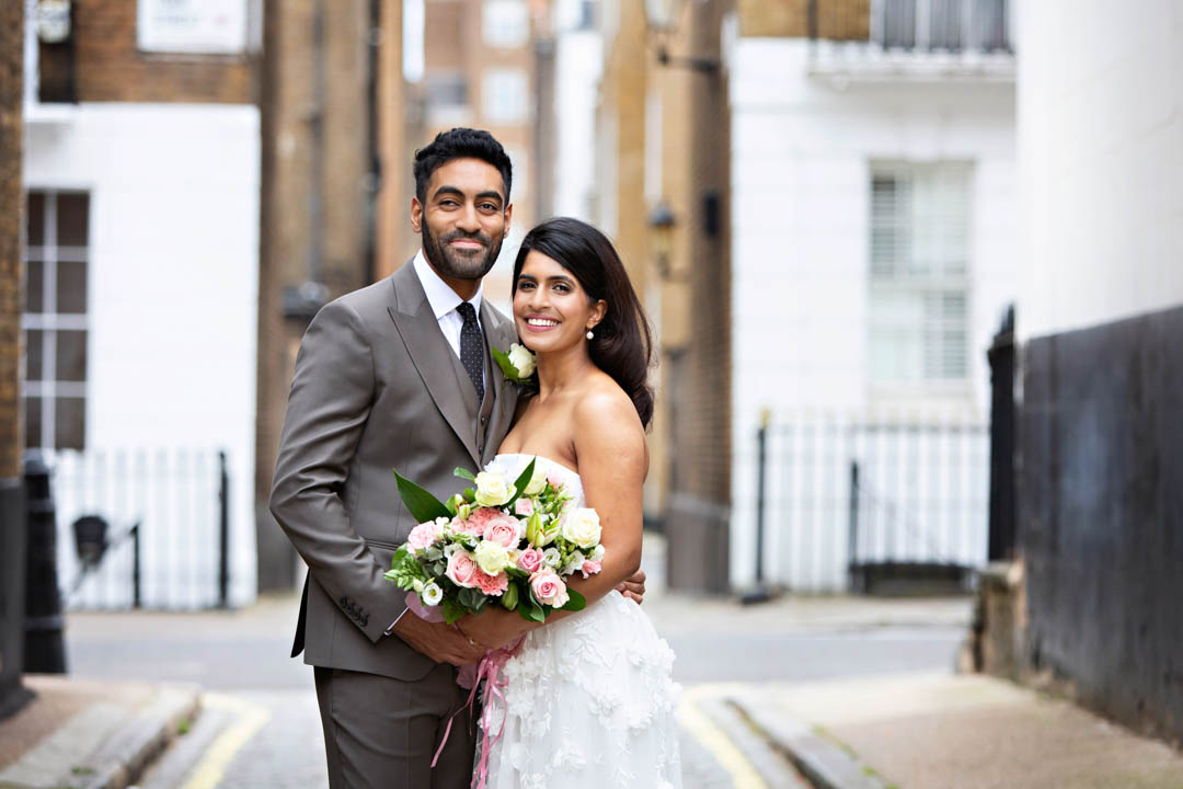 A bride and groom pose for their wedding portrait in a cobbled mews in Marylebone with big smiles on their faces.