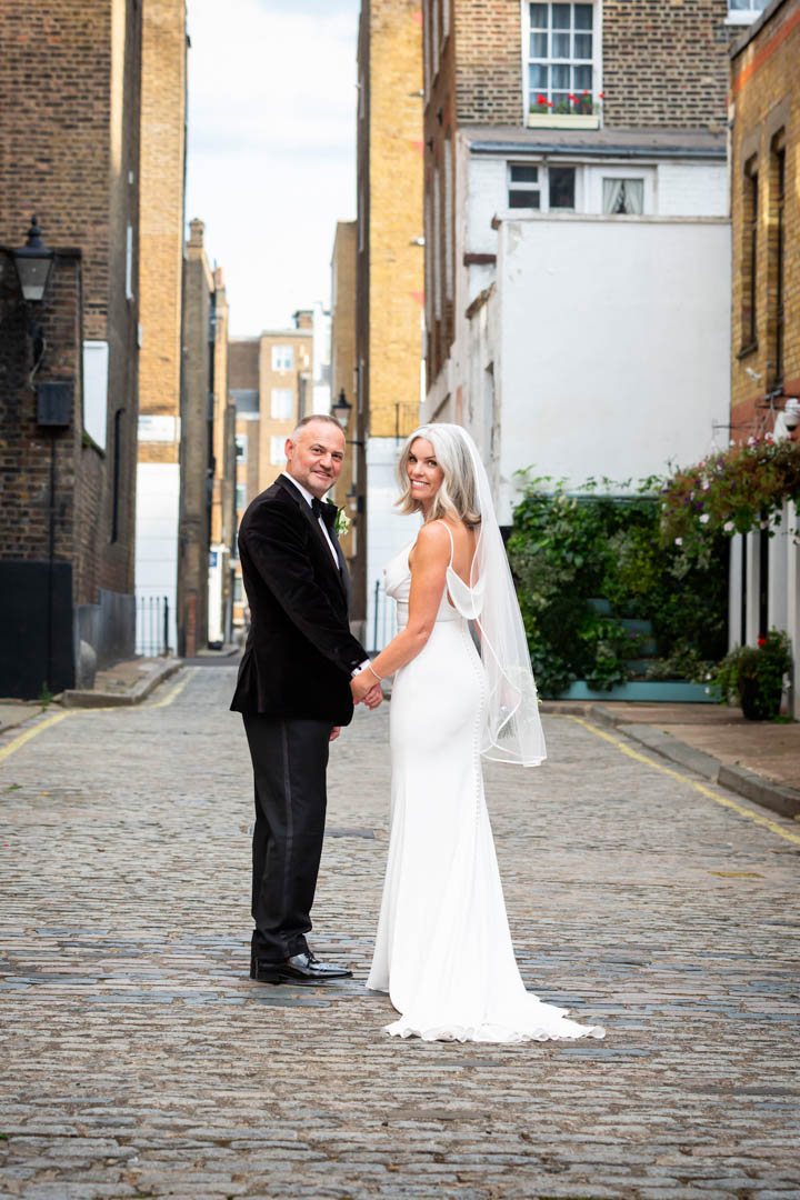 Newlyweds look back the camera with big smiles as they walk down the cobble stoned mews behind The Old Marylebone Town Hall