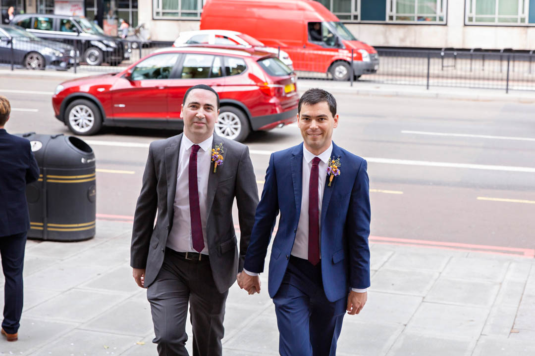 Two grooms hold hands as they walk up the stairs to the Old Marylebone Town Hall together. Their civil partnership ceremony will take place in the Mayfair Room.