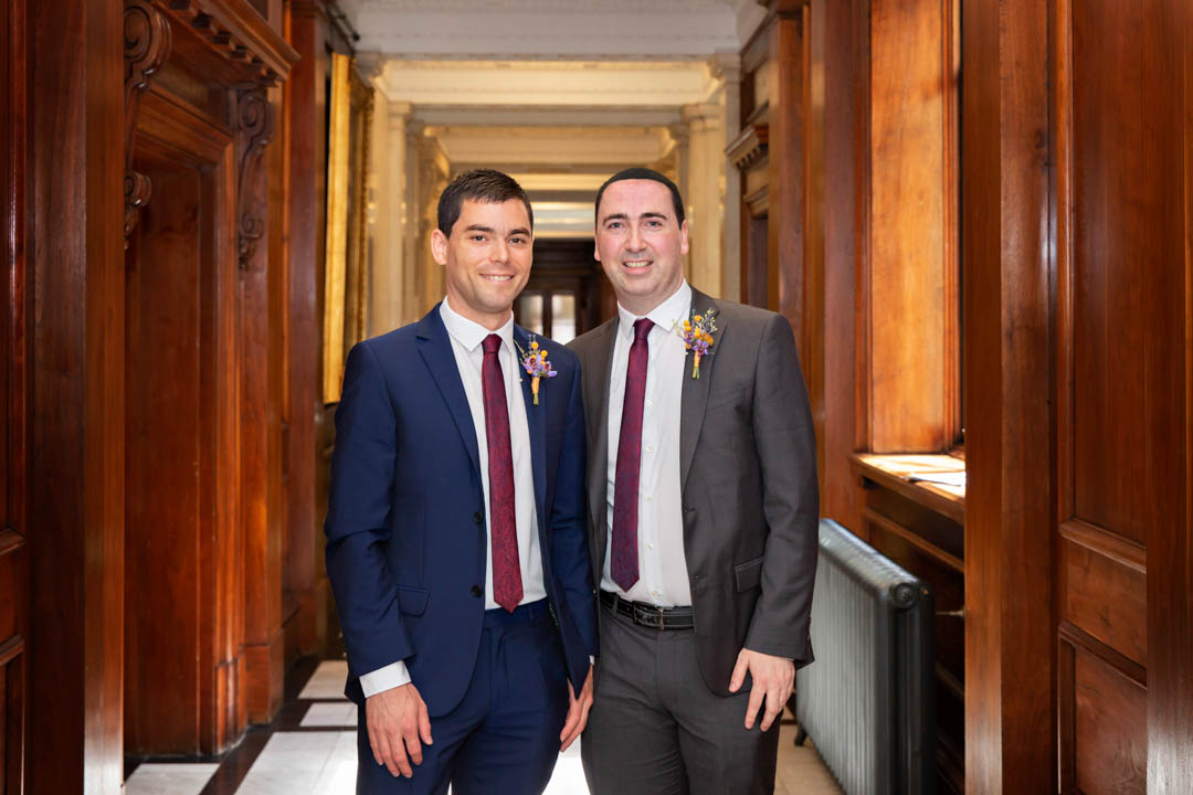 Two grooms stand arm-in-arm in the hallway of the Old Marylebone Town Hall, ready to go into their same sex civil partnership wedding ceremony.