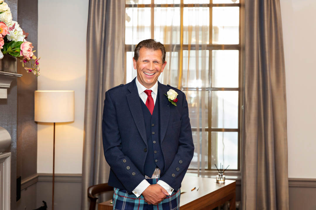 A groom laughs with joy as his bride enters the Mayfair Room wedding ceremony. He is wearing a traditional Scottish wedding suit with tartan trousers instead of a kilt.