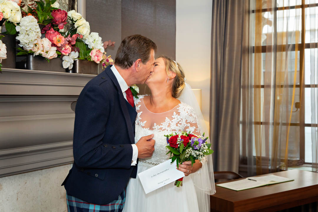 A bride and groom kiss passionately to celebrate the end of their wedding ceremony in the Mayfair Room. The bride is holding their wedding certificate.