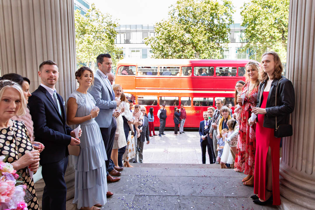 Wedding guests are lined up along the steps of the Old Marylebone Town Hall to throw confetti at the bride and groom. A red vintage London routemaster bus is waiting in the background to take the wedding party to the wedding reception.