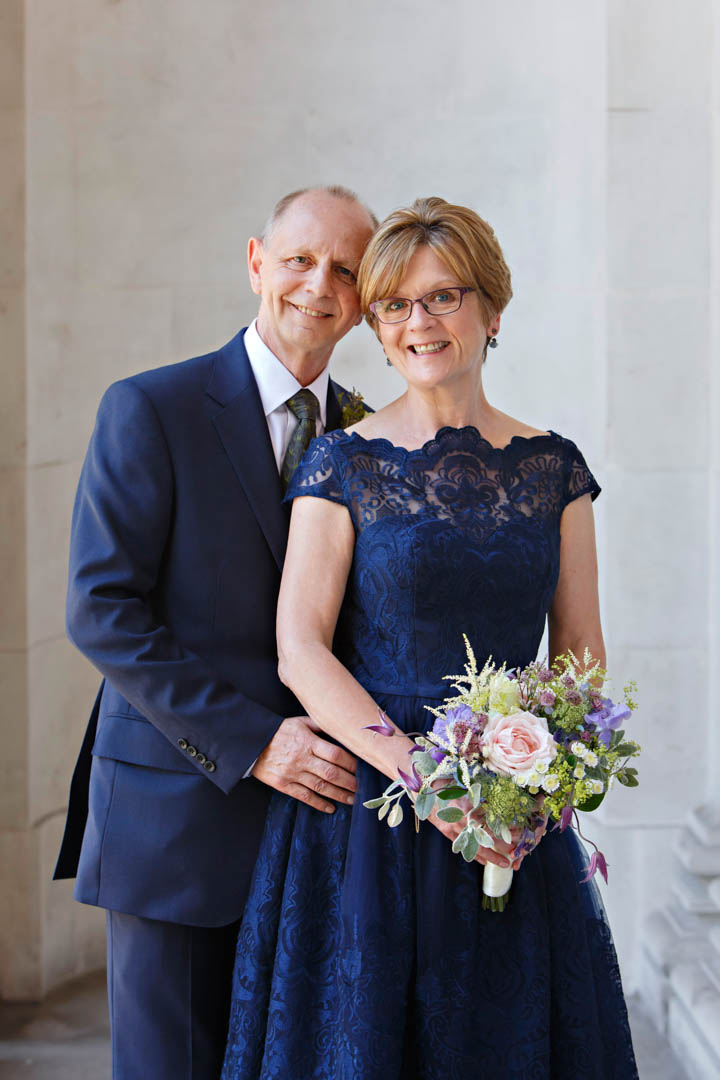 A mature bride and groom pose for a beautifully lit portrait at The Old Marylebone Town Hall. The bride is wearing a blue lace wedding dress.