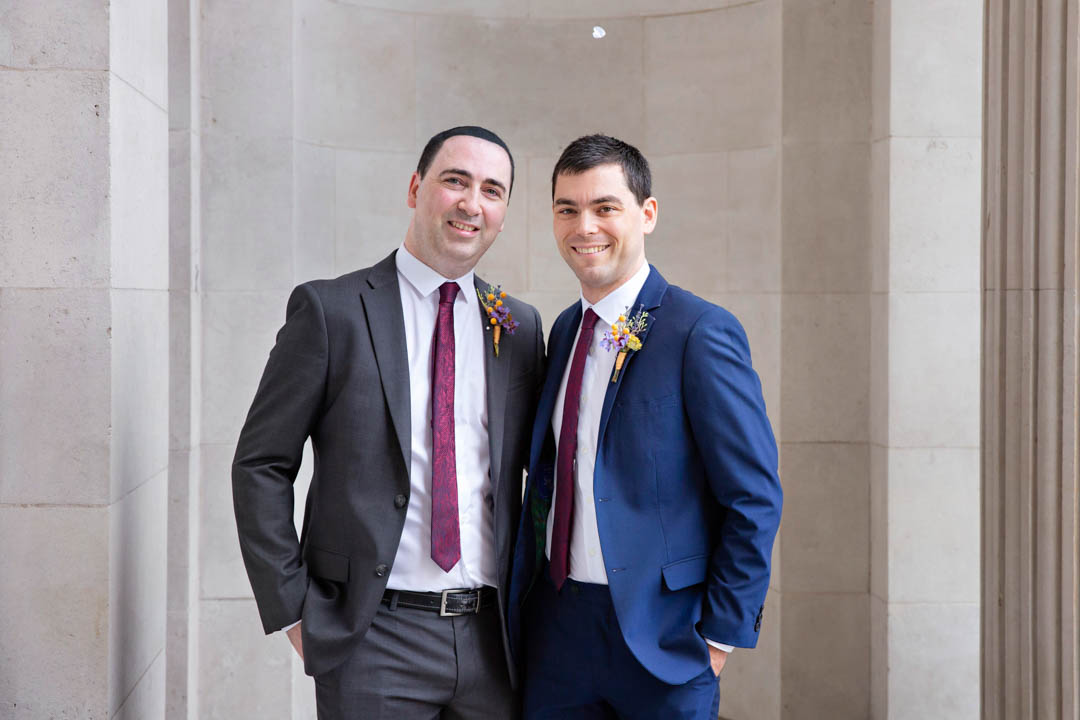 Two grooms pose arm in arm for a same-sex wedding portrait, whilst a single white confetti heart floats in the air above their heads.