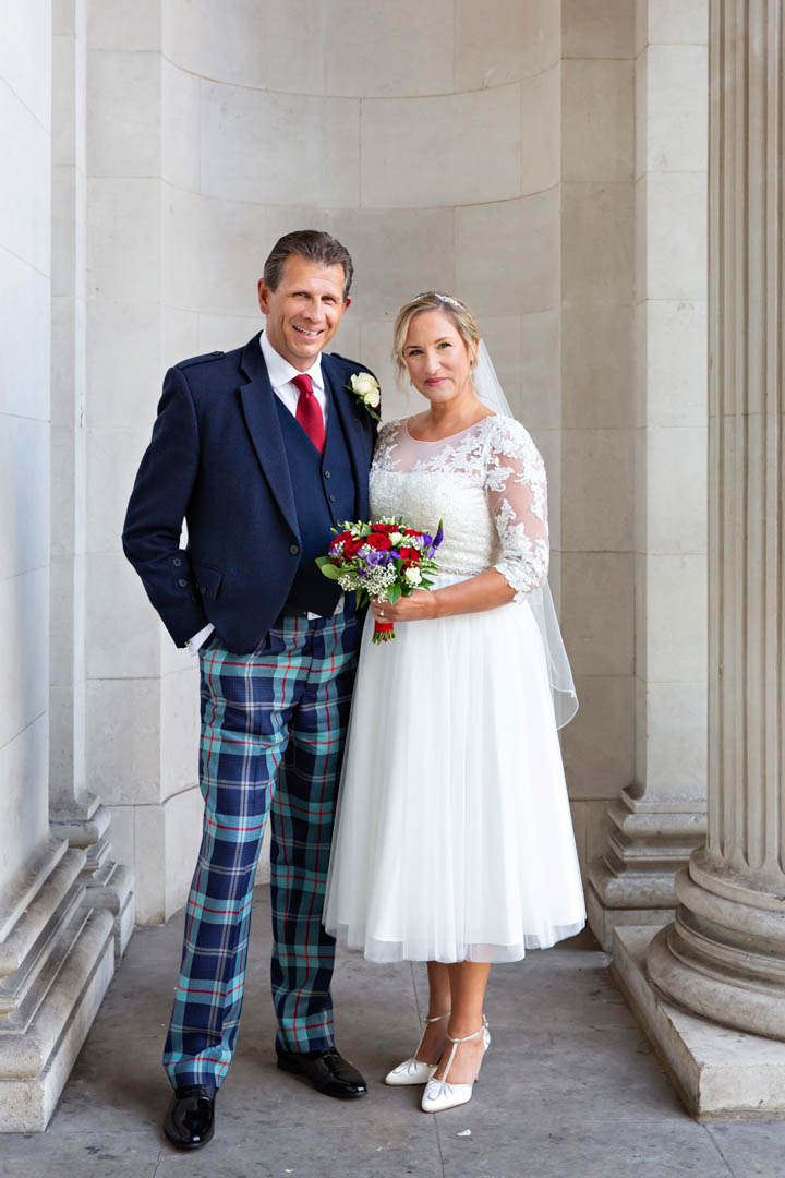 A full-length portrait of a bride and groom following their Mayfair Room wedding ceremony. The bride is wearing a mid-length dress with lace detail on the sleeves. She is wearing a short vail. The groom is wearing a tradition Scottish wedding suit, with tartan trousers.