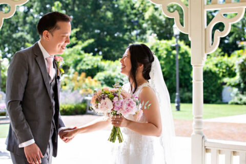 Bride and groom hold hands and smile at each other during their outdoor summer wedding in the gazebo at Morden Park House.