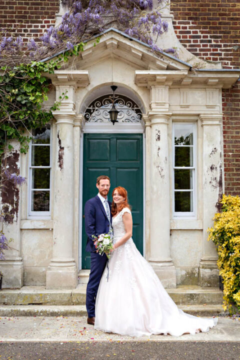 Bride and groom pose for their formal wedding portrait at the entrance to Morden Park House, with Wysteria in full bloom.