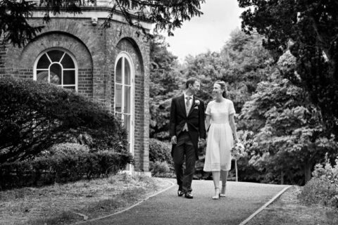 Black and white photograph of bride and groom walking alongside Morden Park House whilst smiling at each other.