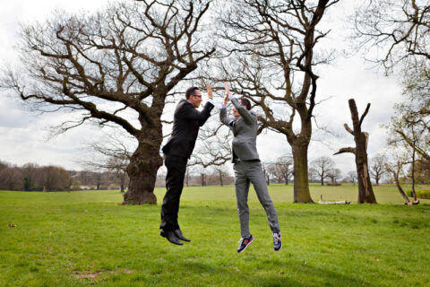 A groom and his best man jump in the air for high-fives to celebrate the groom's marriage.