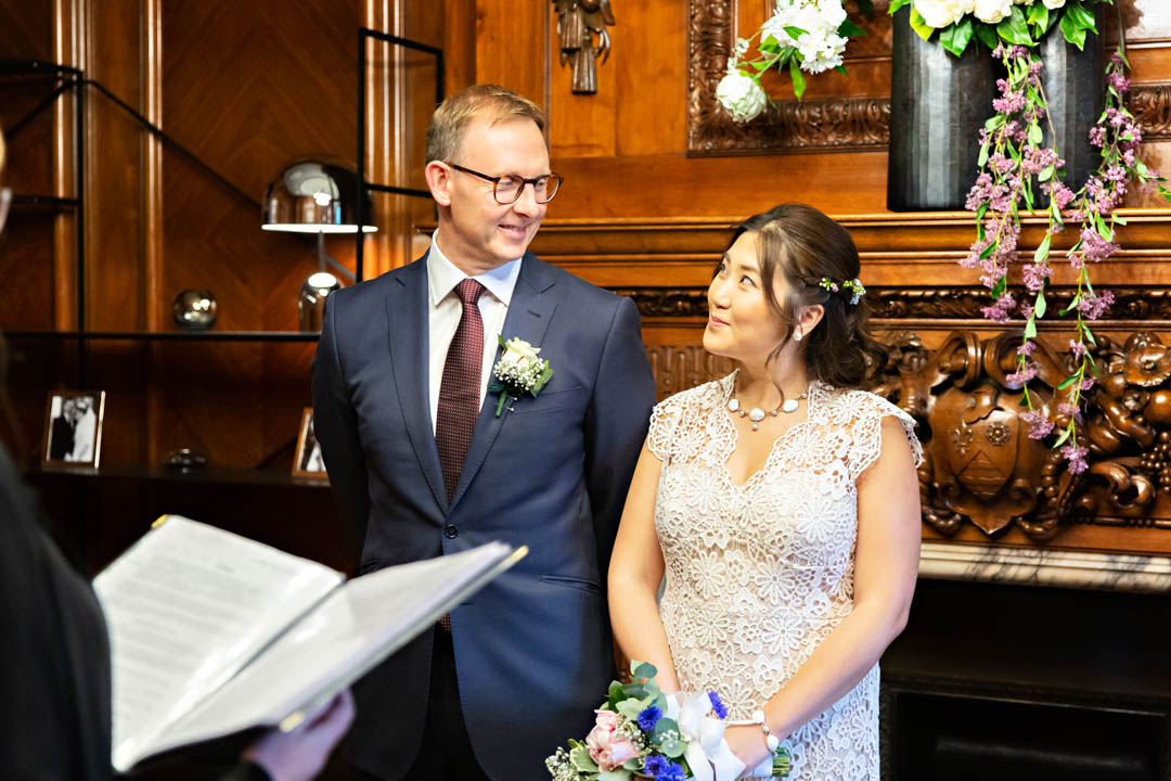 A bride in a lace wedding dress gives her groom a cheeky smile as they exchange wedding vows in the Paddington Room.