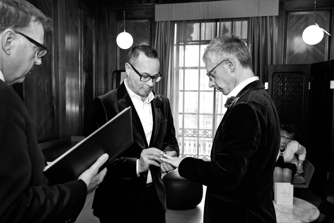 A black and white photo of one groom placing the wedding ring on the second groom's finger during this elopement same sex wedding ceremony in the Paddington Room.