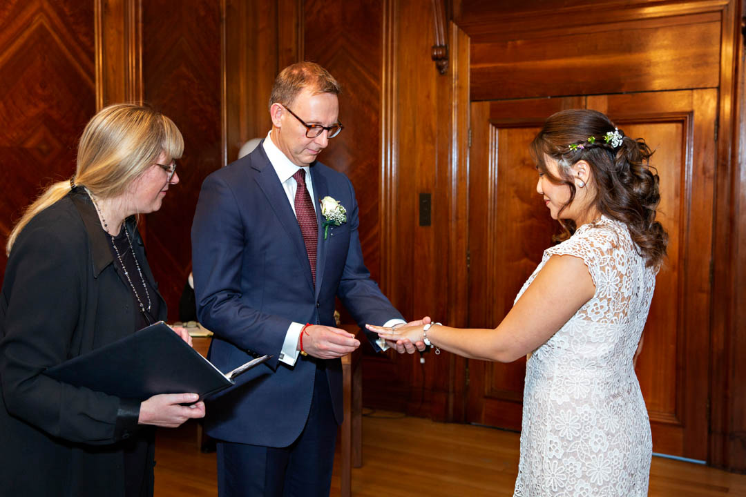 A registrar looks on closely as a groom concentrates on placing the wedding ring on the bride's finger during this register office civil wedding.