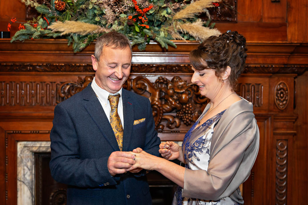 A groom smiles broadly as he places the wedding ring on the bride's ring finger. They are standing in front of the ornately carved wood fireplace in the Paddington Room.