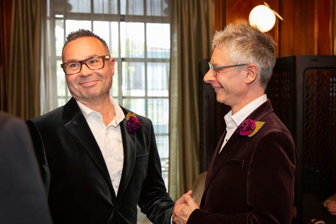 Two grooms hold hands during their same sex wedding ceremony in the Paddington Room. One of the grooms is smiling at the registrar as he is told which words to repeat for his wedding vow.