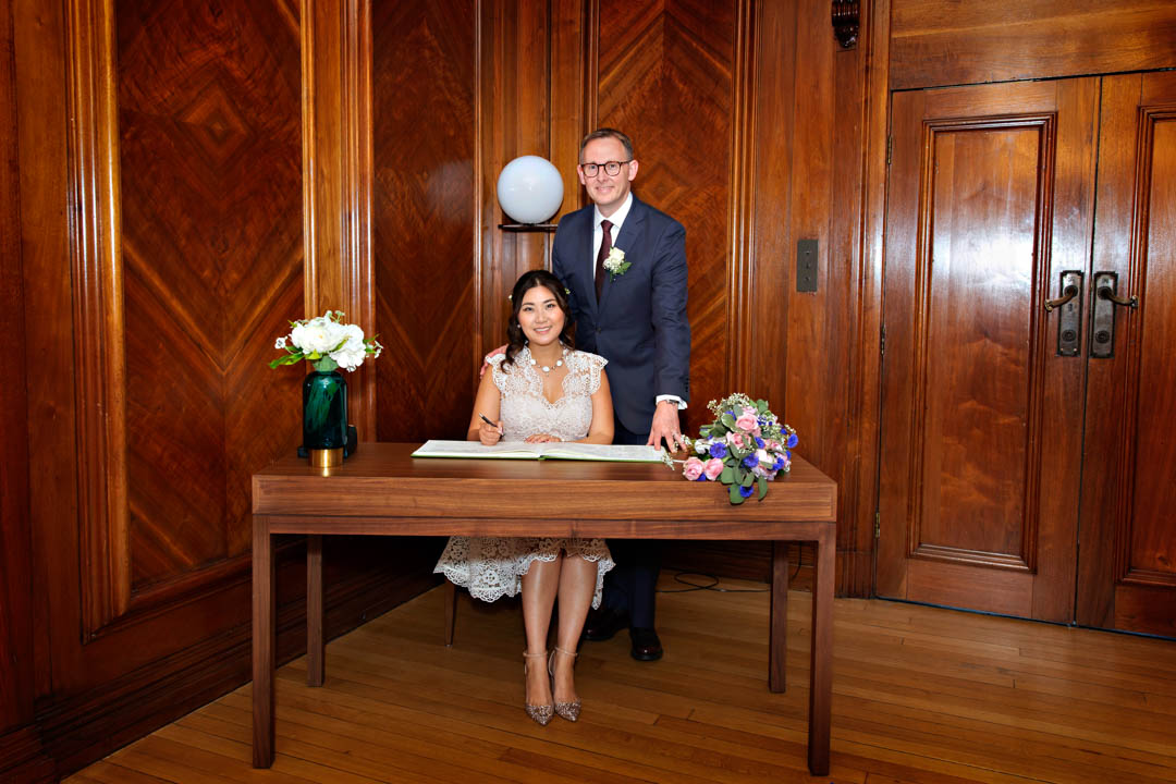 A bride and groom pose with the wedding register book in the timeless wood-panelled Paddington Room.