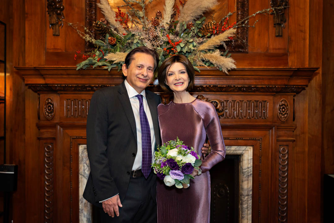 A mature bride wearing an elegant full-length purple dress poses with her mature groom in front of the ornate, wood-carved fireplace of the Paddington Room.
