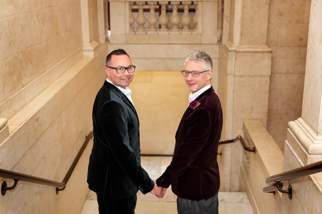Two grooms turn back towards the camera as they walk down the marble steps of the town hall, after getting married in the Paddington Room.