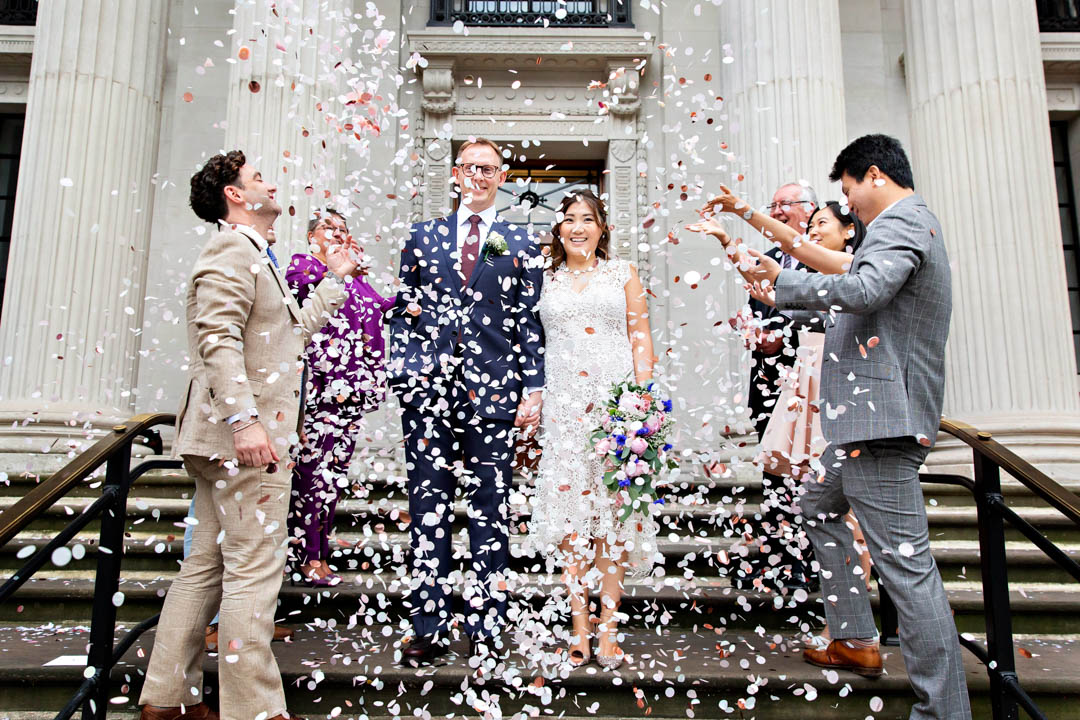 A bride and groom are almost hidden by all the confetti thrown by their guests as they celebrate the end of their register office wedding.