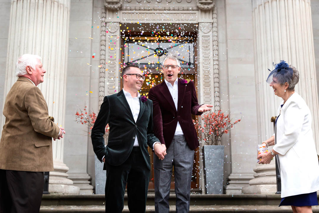 Two grooms exit The Old Marylebone Town Hall into a flurry of colourful confetti.