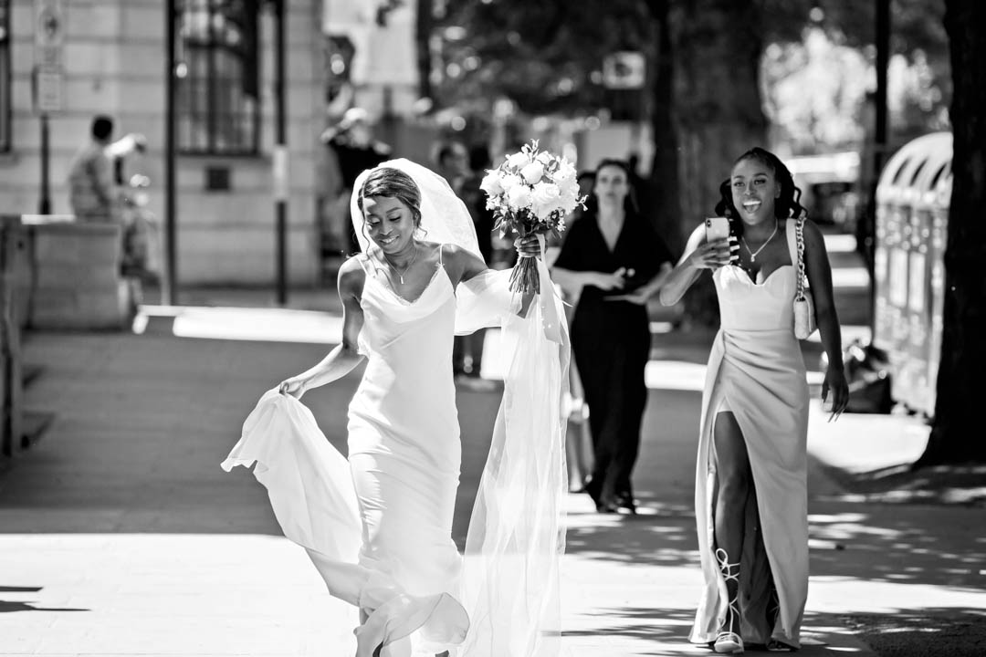 A bride in a white, full-length slip dress holds her train and cathedral length veil aloft as she walks along Marylebone Road on the way to her wedding ceremony in the Pimlico Room in Old Marylebone Town Hall.