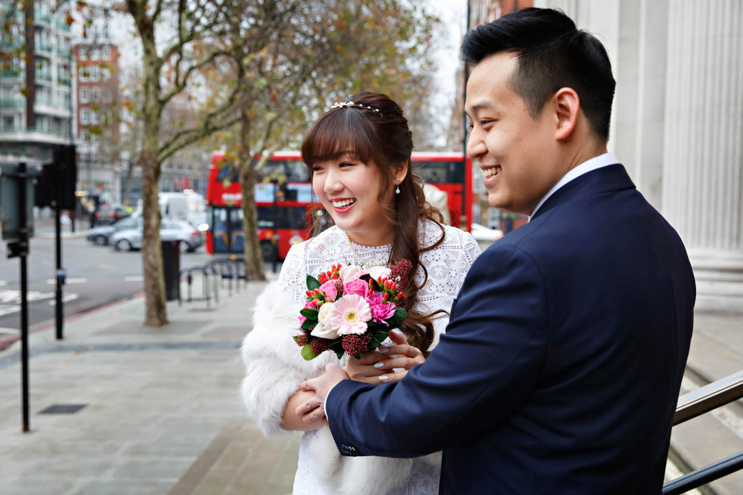 A bride and groom are all smiles on the steps of Old Marylebone Town Hall on Marylebone Road, London. The bride is wearing a white lace dress and holding a red and pink bouquet.