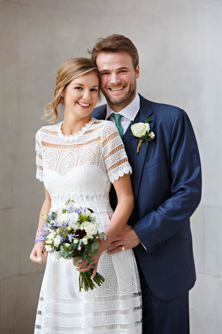 A bride in a white dress and a groom in a blue suit pose for a wedding portrait on the portico of Old Marylebone Town Hall. The bride is holding a mauve and white bouquet.