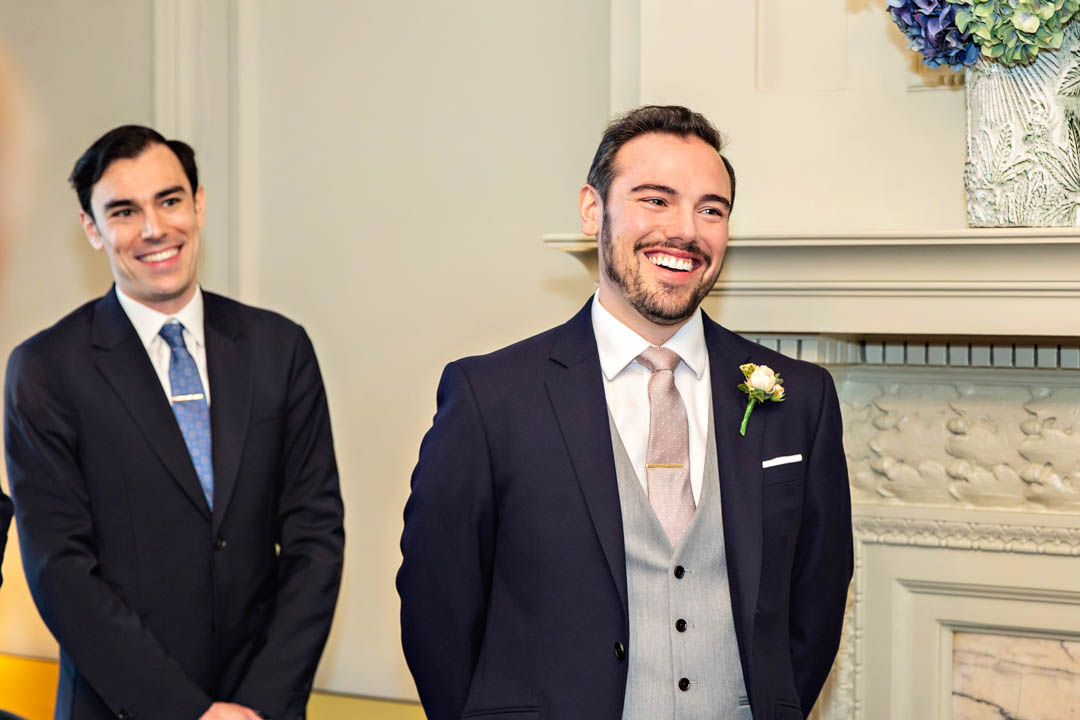 A groom and his best man smile as the bride walks into the Pimlico Room for their wedding.