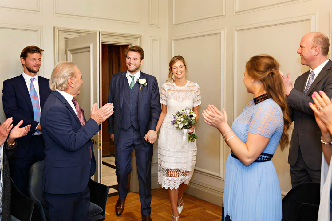A smart groom in a blue three-piece suit and green tie and his bride iin a white lace dress hold hands as they enter the Pimlico Room.