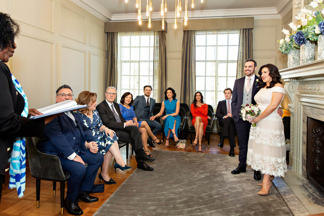 A bride in a white lace midi dress and her groom in a navy suit with grey waistcoat stand together on a grey rug in the Pimlico Room. Their guests sit on chairs on two sides of the room.
