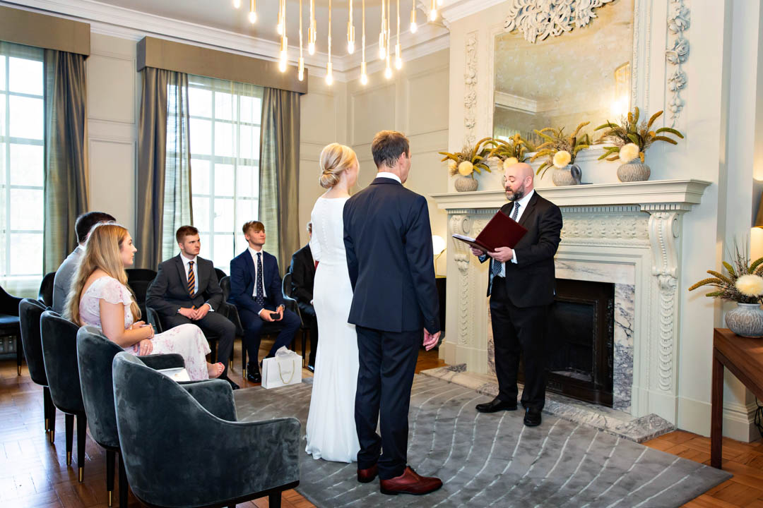 A bride in a full-length white dress and her groom in a dark blue suit stand in front of a Westminster registrar in the Pimlico Room at Old Marylebone Town Hall. They're standing on a blue grey rug in front of their friends and family.