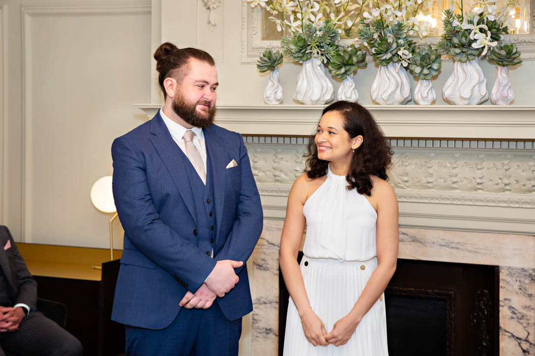 A bride in a halter-necked white dress smiles at her groom, wearing a navy three-piece suit. They're exchanging vows in the Pimlico Room. There are a group of white vases with white flowers on the mantlepiece behind them.
