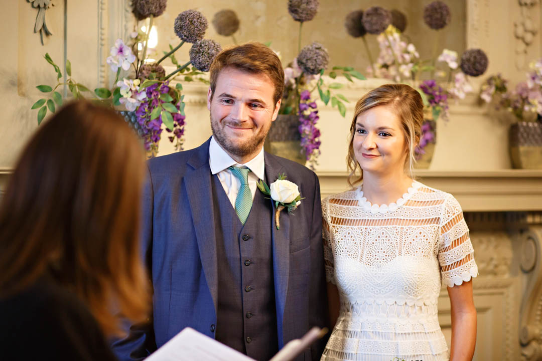 A smiling bride and groom look at a Westminster registrar during their wedding in the Pimlico Room. The groom is wearing a navy three-piece suit and sea green tie and the bride is wearing a white lacy dress.