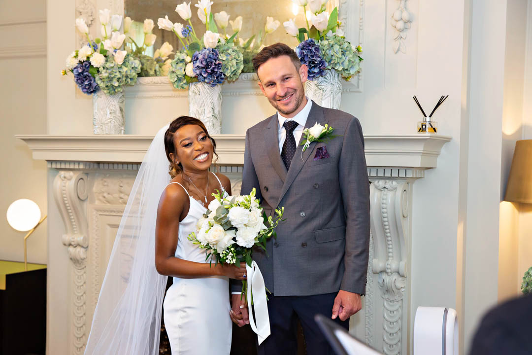 A bride and groom laugh during exchanging vows in the Pimlico Room. The bride is wearing a full-length slip dress and a cathedral length veil. The groom is wearing a double-breasted jacket and dark trousers and tie with a white shirt. There are blue and white flowers in white vases on a mantlepiece in the background.