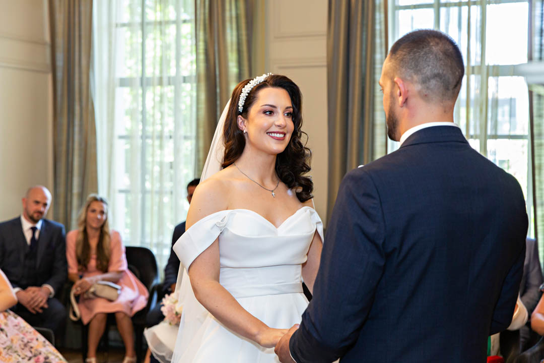 A bride in an off-the-shoulder white dress and white beaded headband smiles as she exchanges vows with her husband in the Pimlico Room. The groom is wearing a navy suit.