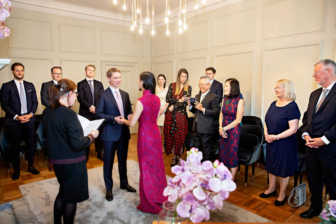 A bride in a full-length pink, patterned cheomsam exchanges vows with her groom in a navy suit. They're standing on a grey rug in the Pimlico Room, surrounded by their wedding guests and a registrar.