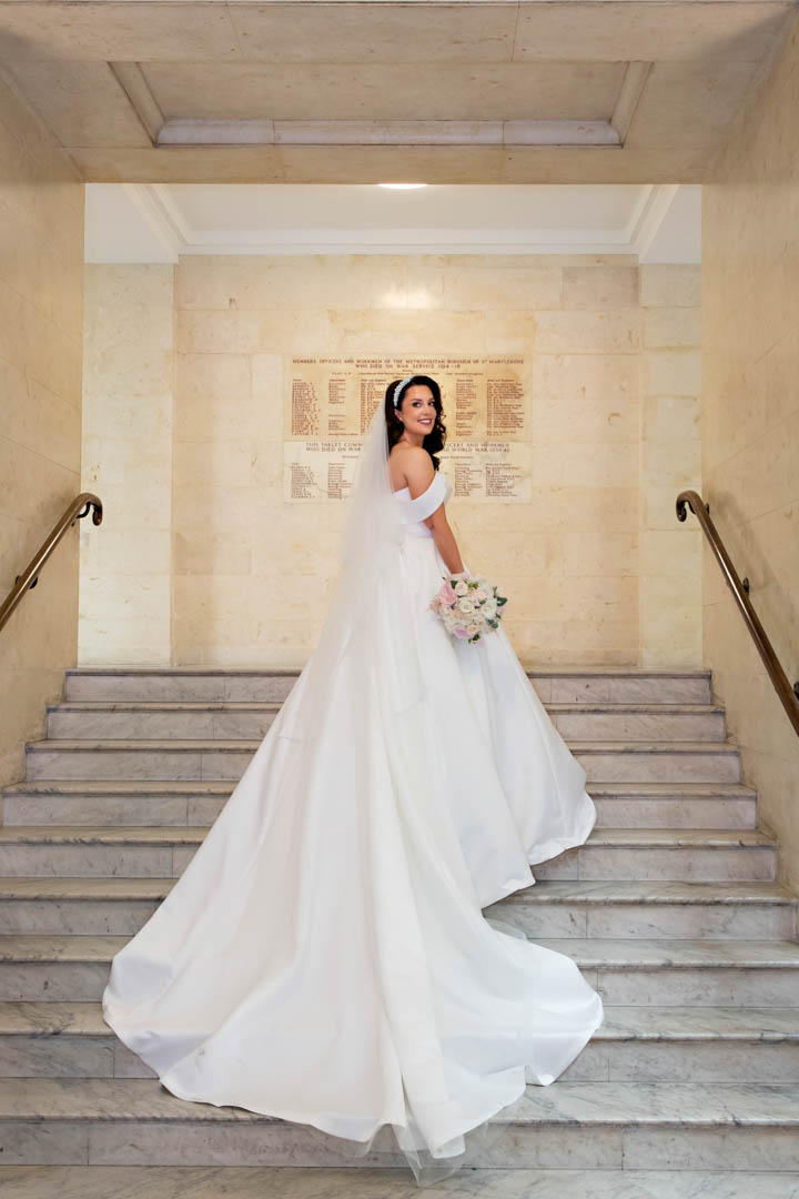 A bride in a full-length white dress with a full train stands on the marble stairs of Old Marylebone Town Hall. The bride is holding a white and bouquet and a white headband.