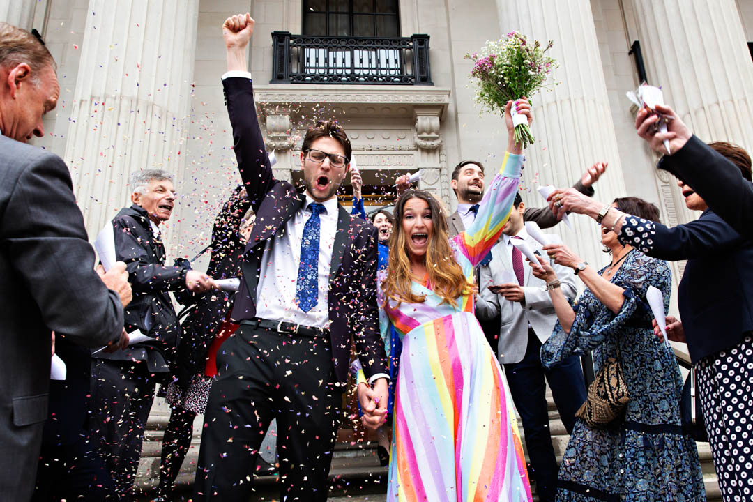 A bride in a rainbow dress and a groom with a colourful blue tie emerge from Old Marylebone Town Hall, after marrying in the Pimlico Room. They're both holding their arms aloft in celebration and the bride is holding a mauve, white and green bouquet.
