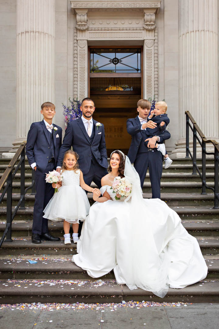 A bride, groom and their four children pose on the steps of Old Marylebone Town Hall. The bride is sitting on the steps.