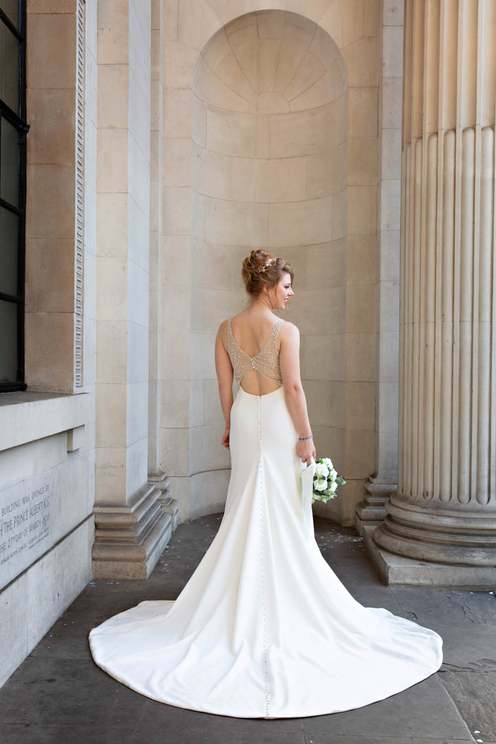 A bride stands with her back to the camera with the train of her white dress in a circle behind her. She's holding a white bouquet.