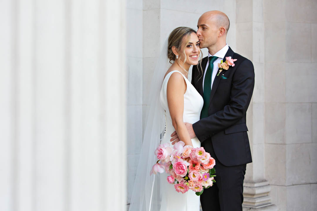 A groom in a dark suit with a dark green tie and colourful buttonhole kisses his bride. She is in a full-length white dress with covered buttons down the back and is holding a spring bouquet in pinks and salmon.