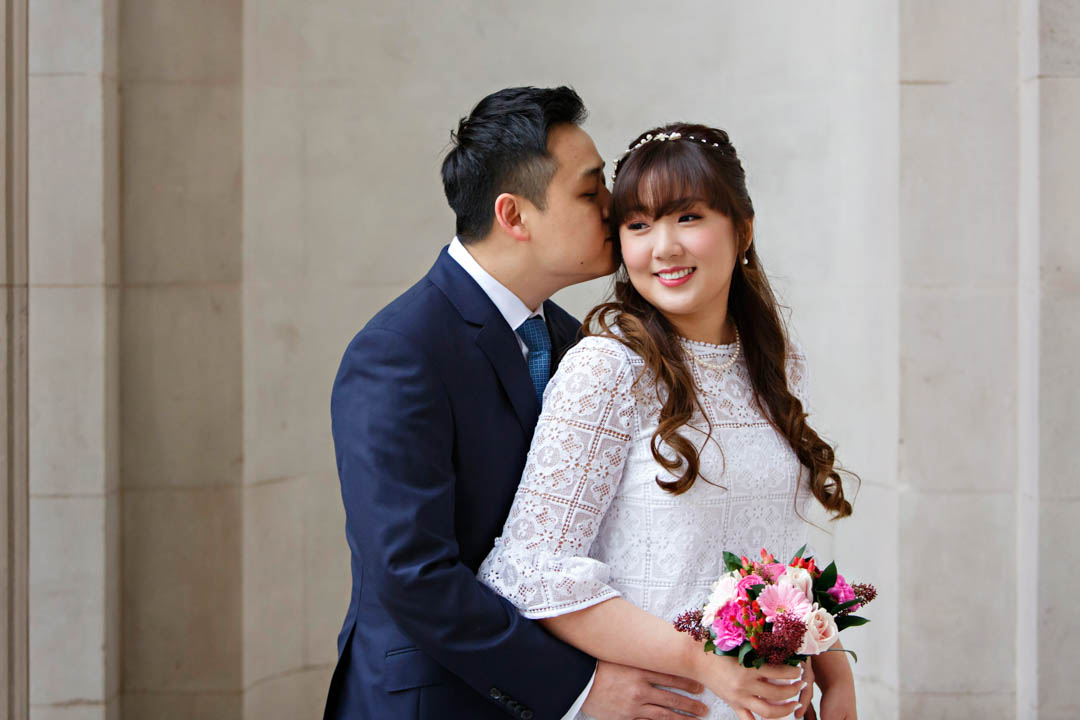 A groom in a navy suit with a mid-blue tie and white shirt kisses his bride on the cheek. She is wearing a white dress with white lace squares and holding a bouquet of pink flowers. They're standing in front of Old Marylebone Town Hall.