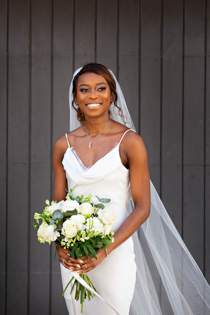 A bride holds a white hand-tied bouquet while posing for a modern bridal portrait against a dark wooden door. She's wearing a white full-length, fitted dress with shoestring straps and a cathedral length veil.