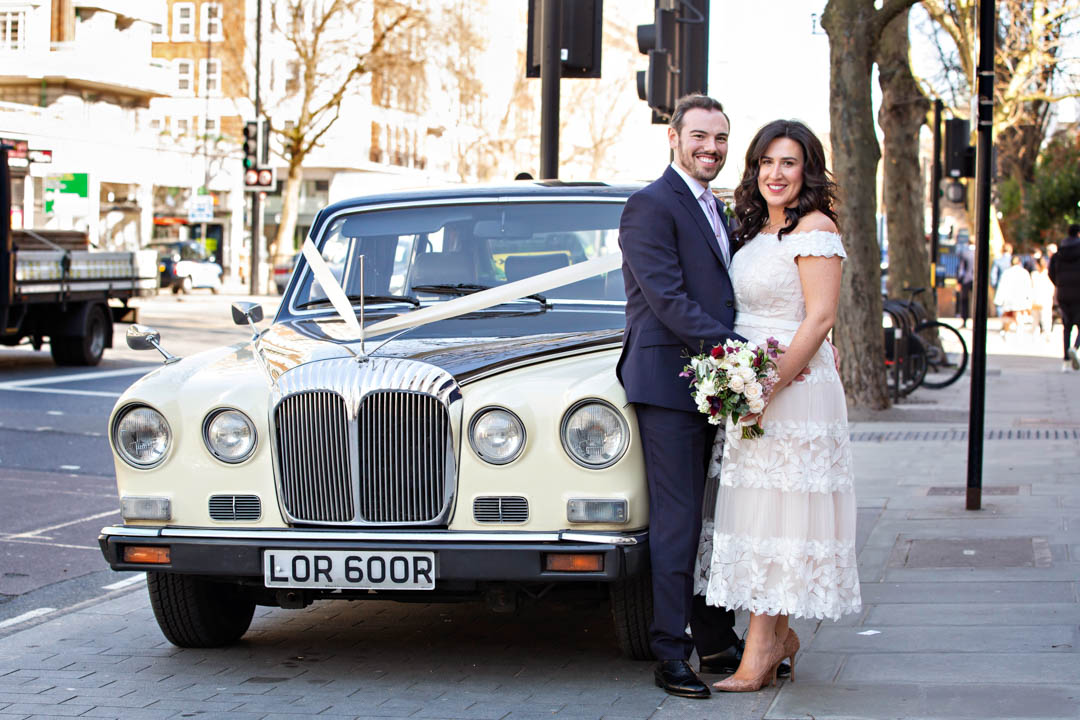A bride in a bare-shouldered lace wedding dress with sheer skirt panels stands next to her groom and a cream Rolls-Royce that is their wedding car for the day.