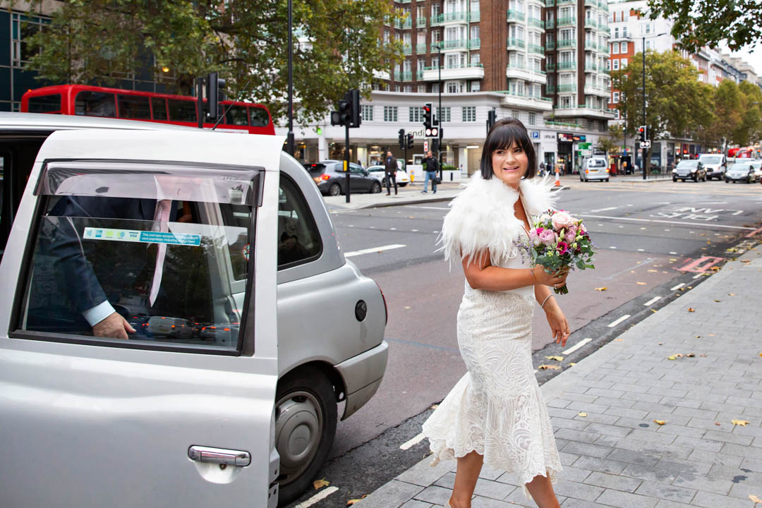 A bride in a fitted white calf-length dress with feathers around her shoulders is holding a pink bouquet. She has just stepped out of a silver London taxi. She's heading to her Soho Room civil marriage ceremony.
