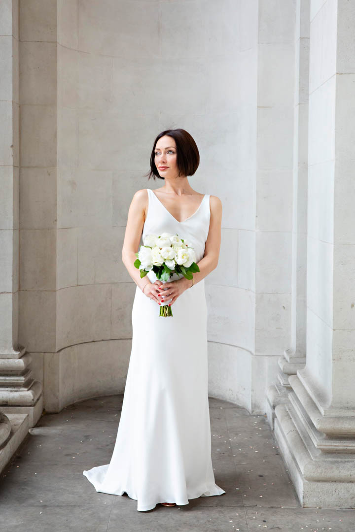 A bride in a sleeveless, v-neck full-length white bridal gown and holding a white rose bouquet stands in the portico of Old Marylebone Town Hall after her Soho Room wedding.