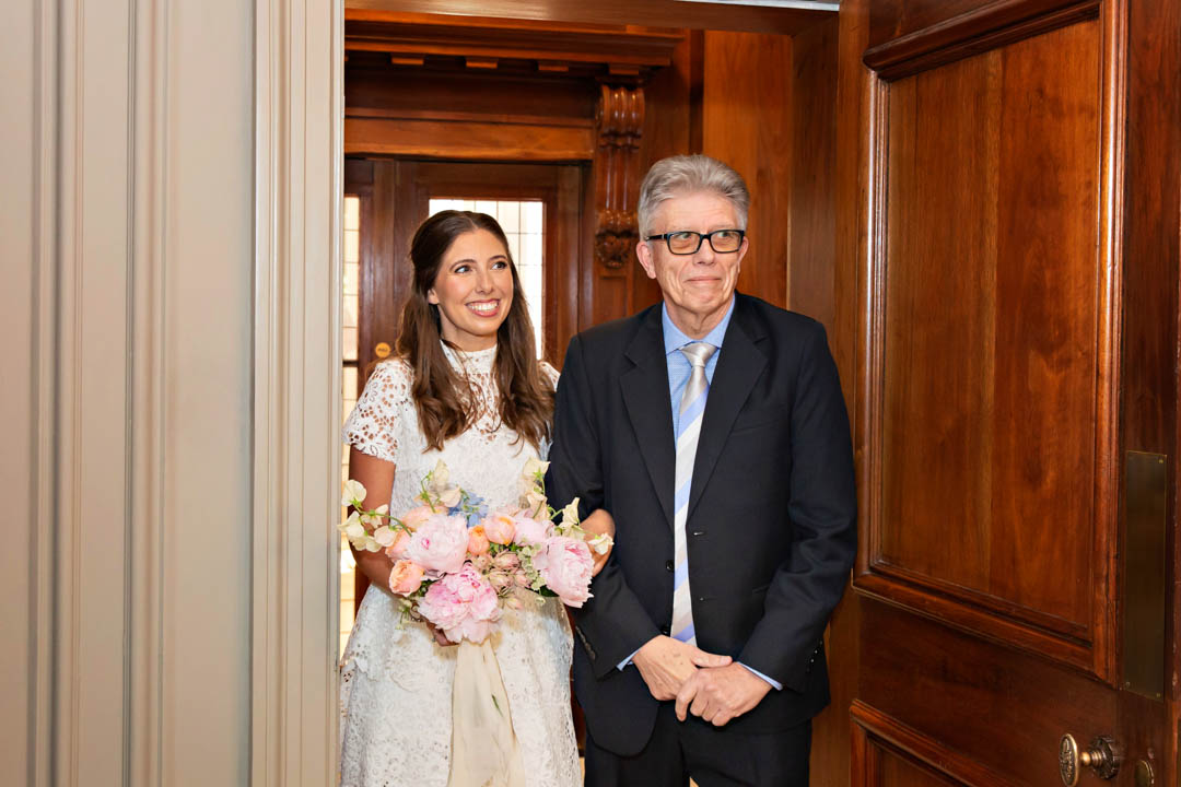 A gentleman in a suit with a pale blue shirt and blue and white striped tie stands in the doorway of the Soho Room, about to walk the bride into the room for her wedding. The bride is wearing a short-sleeved, high necked lace dress and holding a spring bouquet with sweet peas and peonies.